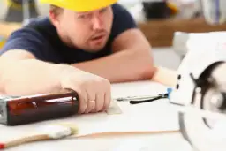 Pracownik w żółtym kasku opiera się o stół, na którym leży butelka i narzędzia.