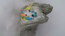 Trzech wspinaczy na skalnym występie, otoczonym mgłą. Jeden z nich, w niebieskim kombinezonie, wisi nad przepaścią, ciesząc się zdobyciem mnich szczyt.