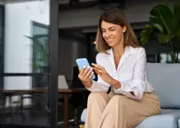 Uśmiechnięta content managerka przegląda coś na telefonie, siedząc na kanapie w nowoczesnym biurze.