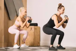 Dwie kobiety wykonujące przysiady z kettlebellami w siłowni.