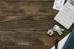 Klucze z breloczkiem w kształcie domu, kalkulator i notatnik na drewnianym tle.