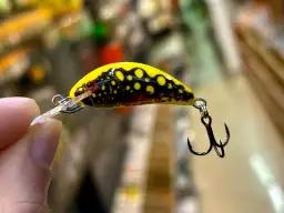 Żółta przynęta wędkarska z czarnymi plamkami i haczykiem, trzymana w palcach.