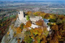 Zamek Chojnik na skalistym wzgórzu, otoczony jesiennymi barwami drzew. W oddali widać miasteczko i pola.