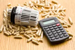 Termostat i kalkulator leżą na drewnie, otoczone przez pellet. To pokazuje, co to jest pellet i jak można obliczyć koszty ogrzewania.