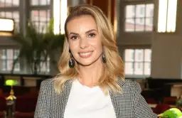 Uśmiechnięta blondynka w marynarce w pepitkę, z dużymi kolczykami. W tle okna i zieleń. Ciekawe, ile lat ma Janachowska?