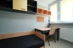 Pokój z łóżkiem, biurkiem i szafkami. Okno z roletami. To wnętrze sugeruje niewielką liczbę izb w domu, idealne dla studenta.