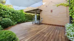 Drewniany taras otoczony zielenią, z dwoma fotelami bujanymi i białym zadaszeniem.