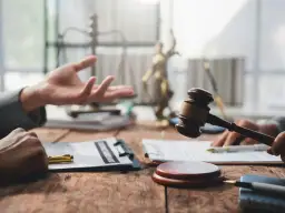 Dwie dłonie przy drewnianym stole, jedna gestykuluje, druga trzyma młotek sędziowski. W tle waga sprawiedliwości i Statua Temidy. Zastanawiasz się, ile się czeka na uzasadnienie wyroku?