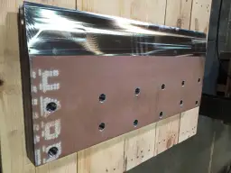 Metalowy element z otworami i oznaczeniami, leżący na drewnianych belkach.