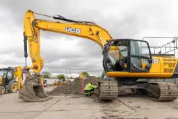 Żółta koparka JCB na placu budowy, pracownik w kamizelce odblaskowej przy gąsienicy.
