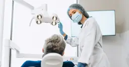Dentystka w masce i rękawiczkach reguluje lampę nad pacjentem siedzącym na fotelu dentystycznym.