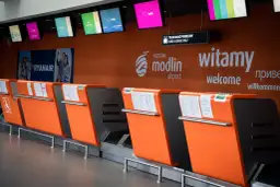 Pomarańczowe stanowiska odprawy bagażowej na lotnisku Modlin z tablicami informacyjnymi i logo lotniska.