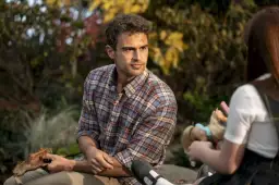 Theo James w kratowanej koszuli, znany z filmów, seriali i programów, siedzi na zewnątrz. Obok niego dziecko bawi się lalką.