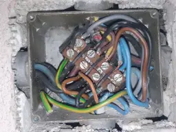 Wnętrze metalowej kostki do kabli z plątaniną przewodów: brązowych, niebieskich, szarych, czarnych i zielono-żółtych, podłączonych do zacisków.