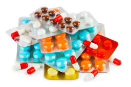 Stos leków w blistrach i kapsułkach: brązowe, białe, niebieskie, pomarańczowe i czerwone.