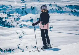 Narciarka w kasku i goglach stoi na stoku, trzymając kijki narciarskie.