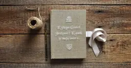 Oto pomysł co zamiast księgi gości na weselu: lniana księga z imionami Justyny i Kamila, ozdobiona wstążką.