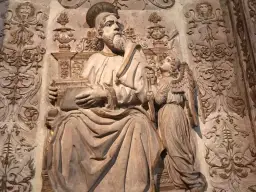 Rzeźba przedstawia św. Mateusza z aniołem, symbolizując znaczenie symboli ewangelistów. W tle widoczne ornamenty.