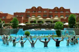 Grupa ludzi trzymających się za ręce w basenie, z hotelowym budynkiem w tle.