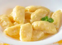 Pyszne, złociste leniwe pierogi w białej miseczce, polane masłem i ozdobione listkiem mięty. Idealne, gdy zastanawiasz się, jak zrobić leniwe pierogi.