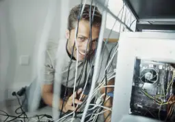 Uśmiechnięty mężczyzna grzebie w plątaninie kabli teleinformatycznych przy komputerze.