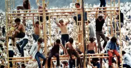 Tłum ludzi na festiwalu, niektórzy wspinają się na konstrukcję. To właśnie woodstock co to za wydarzenie?