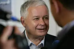 Lech Kaczyński podczas wywiadu, w tle mikrofony i niewyraźne postacie.