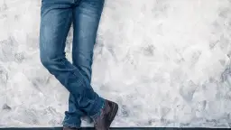 Nogi mężczyzny w dżinsach i brązowych butach stojące przy szarej, teksturowanej ścianie.