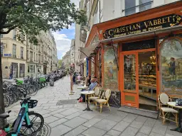 Ludzie siedzą przy stolikach przed cukiernią w Paryżu. Czy 3 dzielnica Paryża jest bezpieczna? Wygląda na spokojną, z rowerami i skuterami zaparkowanymi wzdłuż ulicy.