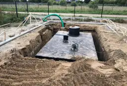 Widok na wykop pod szambo. Duża betonowa płyta, okrągły właz i liczne rury.