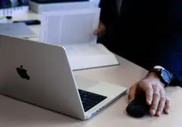 Dłoń mężczyzny w garniturze operuje myszką obok otwartego laptopa i dokumentów.
