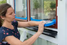Kobieta okleja taśmą malarską drewniane okno, przygotowując je do malowania.
