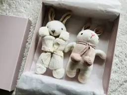 Dwa pluszowe króliczki w pudełku, jeden z kokardką, drugi z szalikiem.