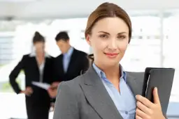 Uśmiechnięta bizneswoman z teczką, w tle współpracownicy omawiający dokumenty. Widać, że panuje między nimi umowa dorozumiana.