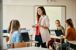 Nauczycielka wyjaśnia studentom, jak zostać nauczycielem po innych studiach. Uczniowie pracują na laptopach w sali lekcyjnej.