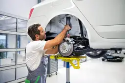 Mechanik naprawia amortyzator w białym samochodzie. To jego praca, a **aso skrót** oznacza, że robi to szybko i sprawnie.