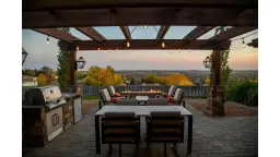 Oświetlenie tarasu inspiracje: przytulny kącik z grillem, stołem i paleniskiem pod drewnianą pergolą, z widokiem na jesienny krajobraz.