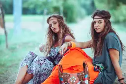 Dwie kobiety w strojach w stylu lat 70. siedzą na trawie, ubrane w kwieciste sukienki i opaski na głowę.