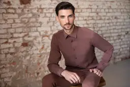 Mężczyzna w brązowej koszuli na wierzchu i spodniach siedzi na tle ceglanej ściany.