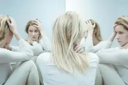 Kobieta z blond włosami w białej bluzce, trzymająca się za głowę, otoczona swoimi odbiciami w lustrze.