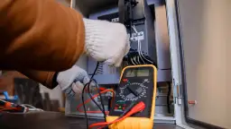Elektryk w rękawiczkach sprawdza połączenia w szafie sterowniczej za pomocą żółtego multimetru. Pokazuje, jak używać multimetru do pomiarów.