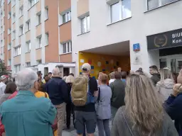 Grupa ludzi stoi przed blokiem mieszkalnym z kolorową ścianą i numerem 2.
