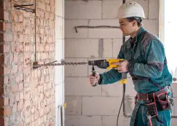 Budowlaniec w kasku pokazuje, jak wiercić w betonie zbrojonym, używając młotowiertarki.