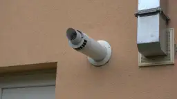Boczny wyrzut spalin przez ścianę budynku z jasną elewacją, obok metalowego kanału wentylacyjnego.