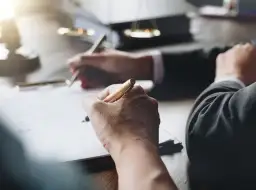 Dłonie piszące długopisami na dokumentach, w tle waga sprawiedliwości.