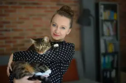 Kobieta w czarnej bluzce w kropki trzyma na rękach pręgowanego kota. To jej profilowe zdjęcie z kotem.