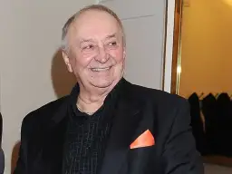 Uśmiechnięty Bohdan Łazuka w czarnym garniturze z pomarańczową poszetką.