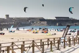 Plaża pełna kolorowych latawców i ludzi, gotowych do **kitesurfingu w Polsce**. W tle widać zabytkowe budowle.