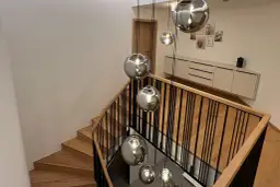 Nowoczesna klatka schodowa z drewnianymi stopniami i czarną balustradą, ozdobiona wiszącymi kulistymi lampami.