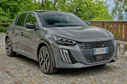 Szary Peugeot 208 zaparkowany na brukowanej drodze, z zielenią w tle.